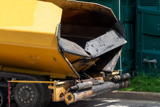 Rear view of a heavy-duty yellow asphalt paver with its hopper open and covered in dark asphalt residue, indicating ongoing road construction and paving work. Industrial machinery details.