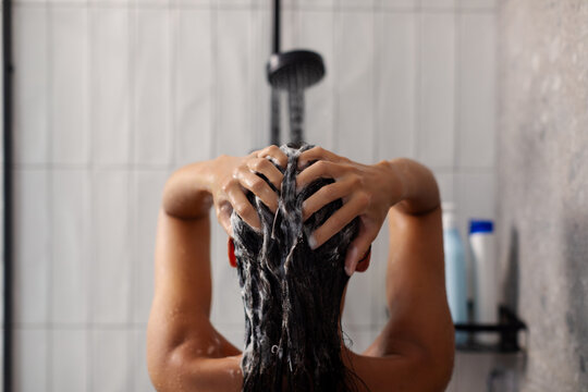 Back view of lady applying shampoo and washing hair under shower, doing haircare routine using cosmetics