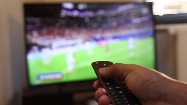 Person changing channels with a remote control while watching a live broadcast of a soccer game, highlighting home entertainment and modern streaming technology