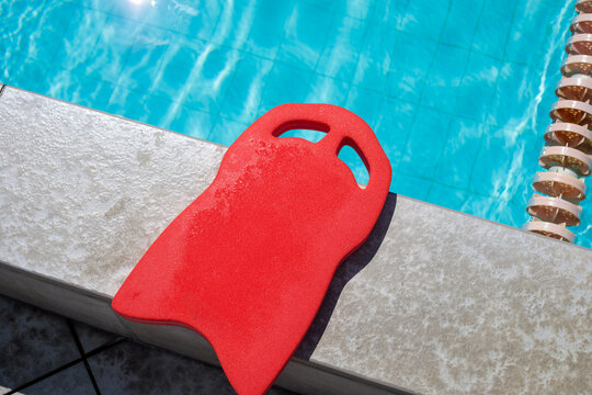 Red swimming kickboard placed on pool coping at edge of outdoor pool, wet kickboard with handle holes resting on stone tile beside lane rope, clear blue pool water with tile bottom visible behind the 