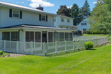 Fototapeta premium Charming white suburban house with a modern metal roof, inviting screened porch, lush green lawn, and classic white picket fence under a sunny blue sky.