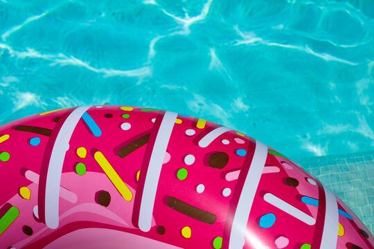 Closeup of pink donut shaped inflatable pool float with white stripe and multicolor sprinkle pattern, donut float top surface in sharp focus with blurred turquoise pool water behind the float in summe