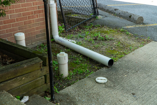 Exterior view of a functional storm drain system featuring white PVC pipes, a cleanout cap, and drainage elements alongside a brick wall, concrete pavement, and overgrown grass in an urban setting.