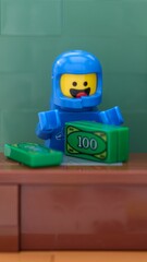 Naklejka premium Tambov, Russian Federation - May 26, 2025 Close-up shot of a blue Lego astronaut minifigure counts green money at a brown wooden desk.