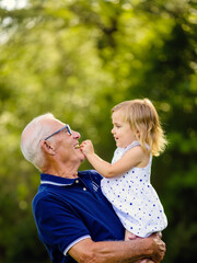 Obraz premium A happy senior grandfather holds his little granddaughter in a sunny garden, sharing a tender moment of laughter and connection during a bright summer day outdoors.