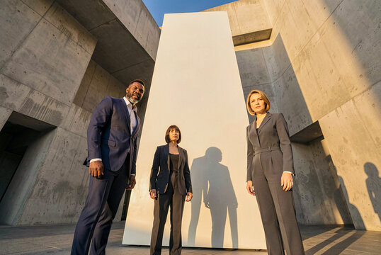 People in formal attire stand before a tall white monolith amidst massive concrete structures under warm golden hour sunlight with sharp shadows cast on vertical surfaces.