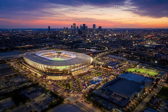 Soccer stadium illuminated at sunset with city skyscraper silhouette. Aerial view of sport arena hosting championship game. Urban night landscape with floodlight during athletic event