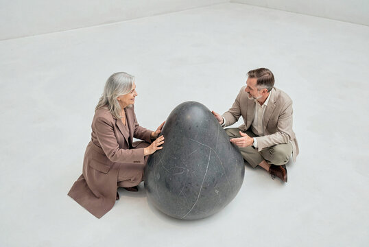 Elderly couple kneels on white floor around a large smooth dark stone sculpture. Overhead bright light casts soft shadows across the minimalist indoor studio space.