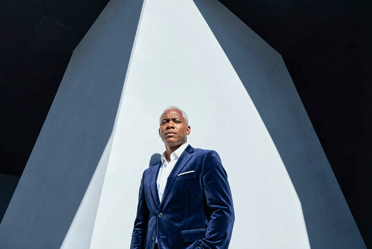 Man with silver hair wears a navy velvet blazer against a sharp geometric concrete structure. Harsh sunlight creates strong diagonal shadows across the white wall behind him.