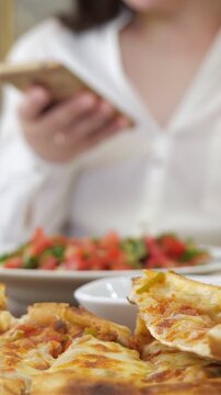 Delicious traditional turkish pide in the foreground with a woman out of focus using her smartphone and a fresh tomato salad in the background, representing a modern dining experience