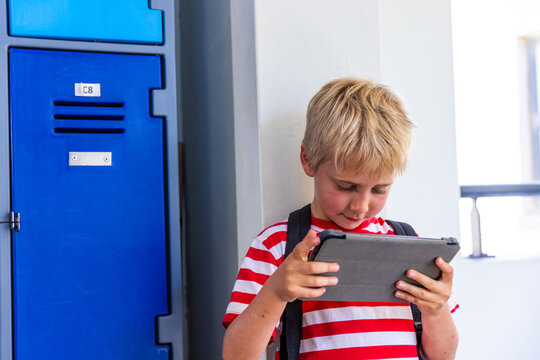 School-age boy standing in school hallway by blue locker labeled C8, holding grey tablet