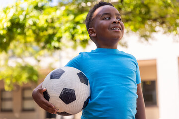 Naklejka premium African American school-age boy wearing blue T-shirt holding soccer ball and smiling in schoolyard