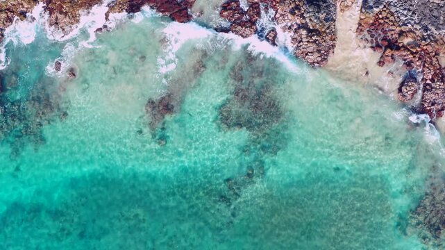 Overhead view of ocean waves rolling toward the shore, revealing shifting shades of green and turquoise with bright white foam patterns. The dynamic movement of the water creates a powerful Hawaii