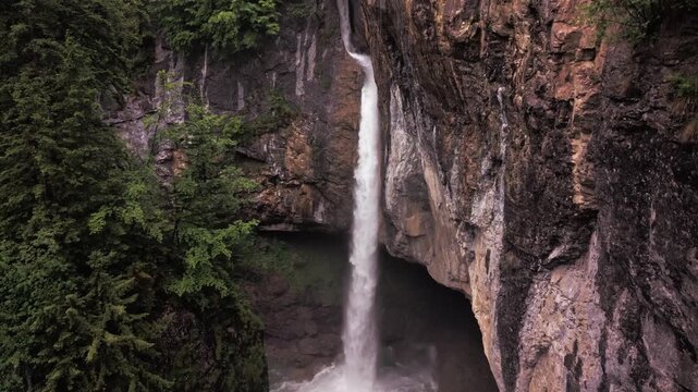 Aerial view of tall mountain waterfall plunging into dark rocky gorge surrounded by trees