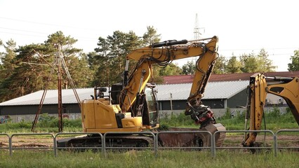 Żółta koparka gąsienicowa z widocznym ramieniem roboczym na placu budowy. Maszyna stoi za metalowym ogrodzeniem, a w tle widać budynek, zielone drzewa oraz jasne niebo. © tomo81