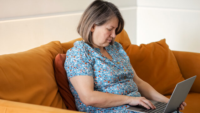 Individual Focused On Laptop In Peaceful Setting. Female Student Concentrating On Course Material At Home. Woman Sitting Comfortably On Sofa While Attentively Studying And Reviewing Notes Online
