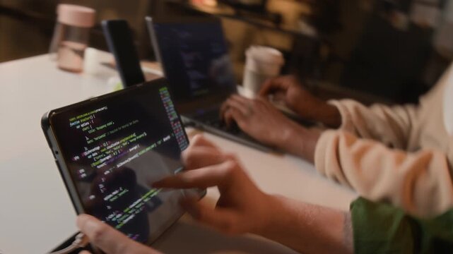 Two unknown young programmers writing and reviewing code on tablet and laptop in office. Video for software development, IT startups, and tech education marketing