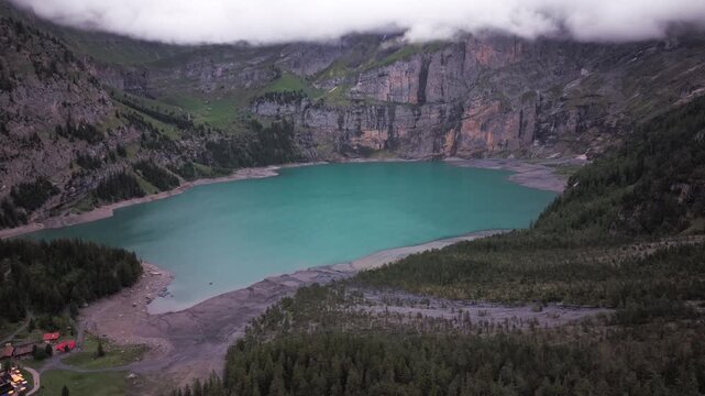High-angle aerial panorama of a vibrant blue mountain lake nestled beneath massive rocky cliffs and dramatic low clouds. Captures the majestic, moody wilderness of the European Alps.