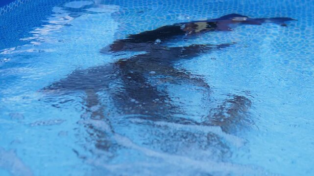 An adult swimmer moves beneath the rippling surface of a bright blue swimming pool, viewed from above with clear water, mosaic tiles, and shimmering sunlight creating a calm