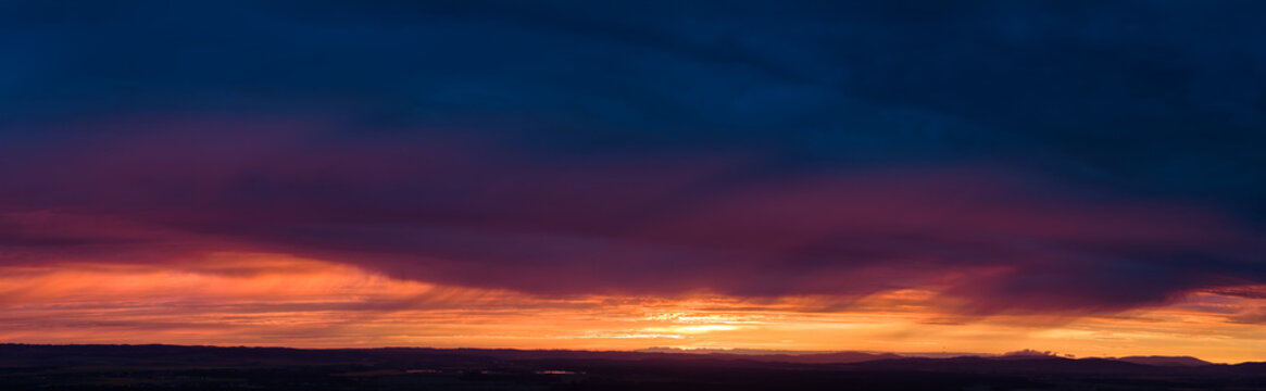 Panoramic,  dramatic sunset sky with dark cloud layers and a glowing orange horizon, ideal as a wide natural background. Colors: blue, orange, black.