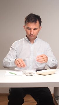 Vertical video man holding an empty wallet at a white table, contemplating his financial situation with a notepad, pen, scattered money, and a calculator nearby