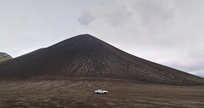 White pickup car truck traversing the expansive ash plain and volcanic landscape of Port Resolution on Tanna Island, with Mount Yasur's peak dominant against a cloudy sky. Trekking drone flight