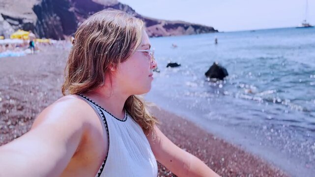 Woman vlogger waving and taking a selfie on red beach in santorini, greece