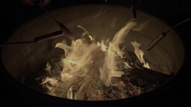 Close-up video of orange campfire flames dancing over glowing coals while hot dogs roast on sticks above the fire pit at night. Outdoor camping and recreation concept.