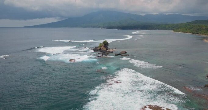 Tanna Island coastline showing a small rocky island with a tree, breaking ocean waves, and green mountains meeting the sea under a dramatic cloudy sky near Port Resolution. Aerial view drone flight