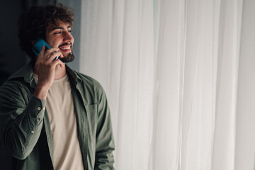 Young man talking on smartphone, smiling happily indoors, communicating remotely with friends or...