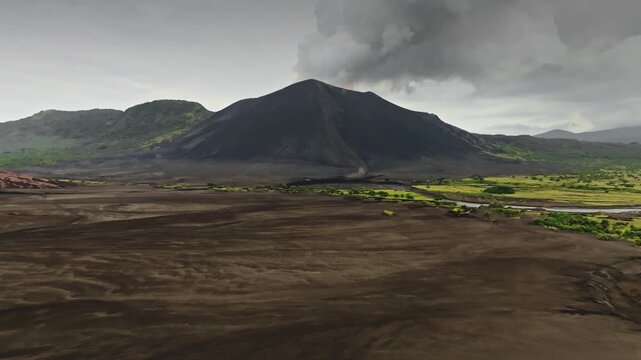 Mount Yasur erupting, ash and smoke plume towering over dark volcanic plain, with winding green river and vegetation on Tanna Island, Vanuatu, under cloudy dramatic sky. Aerial view drone flight