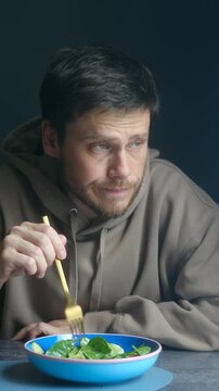 Unhappy man sitting at table picking fresh vegetable salad with fork, making dissatisfied face. He does not want to eat boring tasteless salad while on strict diet. Vertical video. Diet concept.