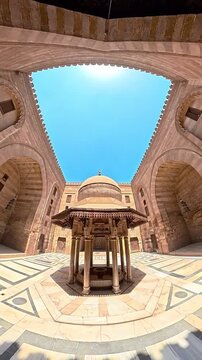 A cinematic wide-angle shot capturing the central ablution fountain (Sabil) within the serene courtyard of the historic Al-Hakim bi-Amr Allah Mosque on Al-Muizz Street, Islamic Cairo, Egypt. The foota