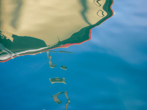 Abstract close-up of a white boat hull reflection distorted by ripples in turquoise-blue marina water, Budva, Montenegro. Fluid painterly shapes in teal, cream and deep blue. Creative abstract