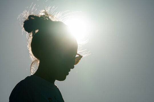 Silhouette of a woman in sunglasses with a messy bun, backlit by bright sun against a clear sky, creating a dramatic summer profile.