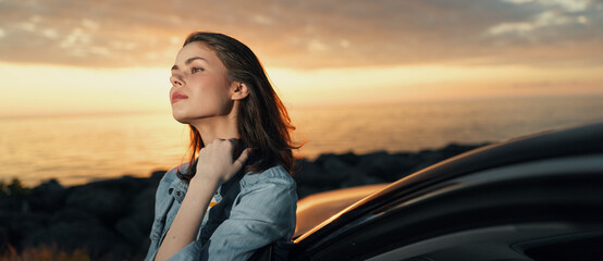 A young woman stands beside a car at golden hour, looking toward the horizon with a calm, contemplative expression beside the glowing sea. © SHOTPRIME STUDIO