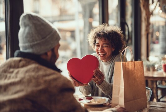 A smiling woman holding a red heart-shaped gift box while sitting at a restaurant table with a man in a beanie during a romantic date night