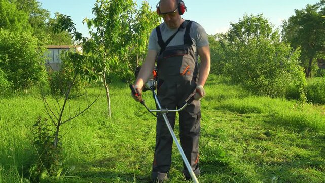 Worker in protective gear uses a grass trimmer to cut grass in a lush garden, surrounded by trees and greenery under bright sunlight