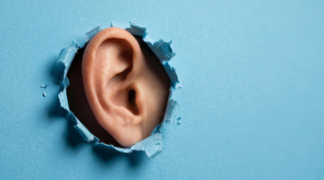 Human ear peeking through ripped blue paper hole showing detail of skin texture and auditory organ for medical concept.