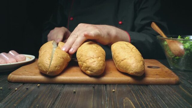 Chef in black attire skillfully slices open bread rolls for hot dogs on a wooden cutting board, with sausages on a plate and a bowl of fresh salad in the background