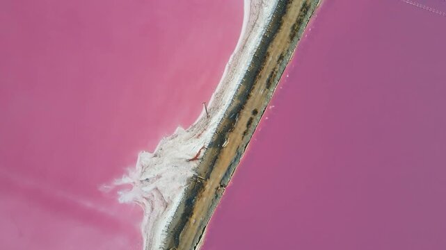 4K drone video capturing narrow dirt road or dam cutting straight through a vibrant pink salt lake. The abstract geometric composition divides the frame into two colorful mineral ponds.
