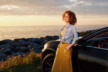 Fototapeta premium Woman lifestyle portrait by a car on a coastal sunset scene, wearing denim jacket and yellow dress, relaxed pose, natural light, calm sea breeze, cinematic warm tones.