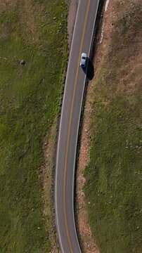 Vertical Screen: Beartooth Highway in Montana USA with silver car traveling along curved mountain road surrounded by green grass patches and snow remnants on arid terrain.