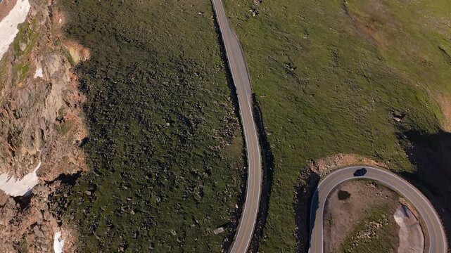 Aerial of Beartooth Highway, Montana, USA showing dramatic winding mountain road with sharp hairpin turn. Drone footage captures vehicle navigating alpine terrain with green tundra and snow patches.