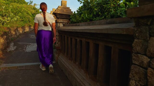 Young Woman Walking Along Historic Stone Temple Path at Pura Luhur Uluwatu During Warm Evening Light