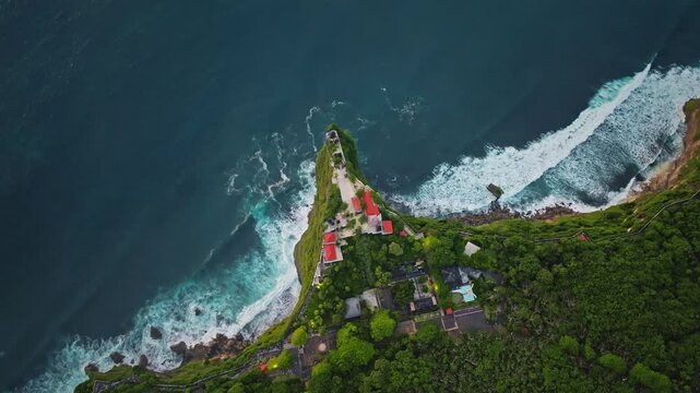 Uluwatu Temple, a revered Hindu sea temple, sits dramatically on a high cliff overlooking the vibrant blue Indian Ocean and crashing waves surrounding Bali's Bukit Peninsula. Aerial view drone shot