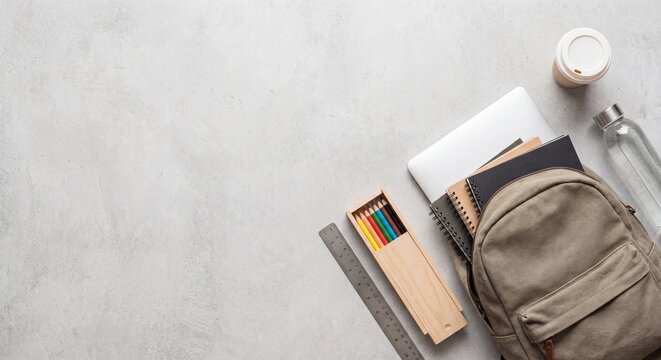 Top view of a brown school backpack next to a laptop colored pencils in a wooden case and a coffee cup on a grey surface