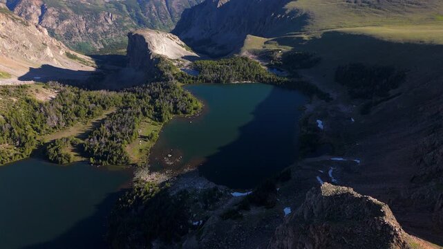 Aerial view of Beartooth Highway, Montana, USA showcases pristine alpine lakes surrounded by rugged mountain terrain, dense evergreen forests, and dramatic rocky peaks under warm golden morning light.