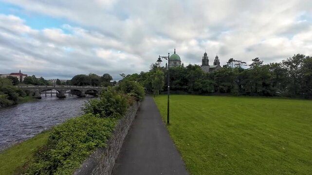 Cinematic aerial drone footage capturing path and green meadow, Galway Cathedral, Corrib River flow and Salmon Weir Bridge in Ireland