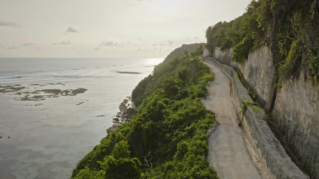 Cliffside road carved in limestone rock hugging the coastline of Bali, Indonesia, with lush green vegetation leading to the ocean and Green Bowl Beach during sunset. Aerial view drone flight